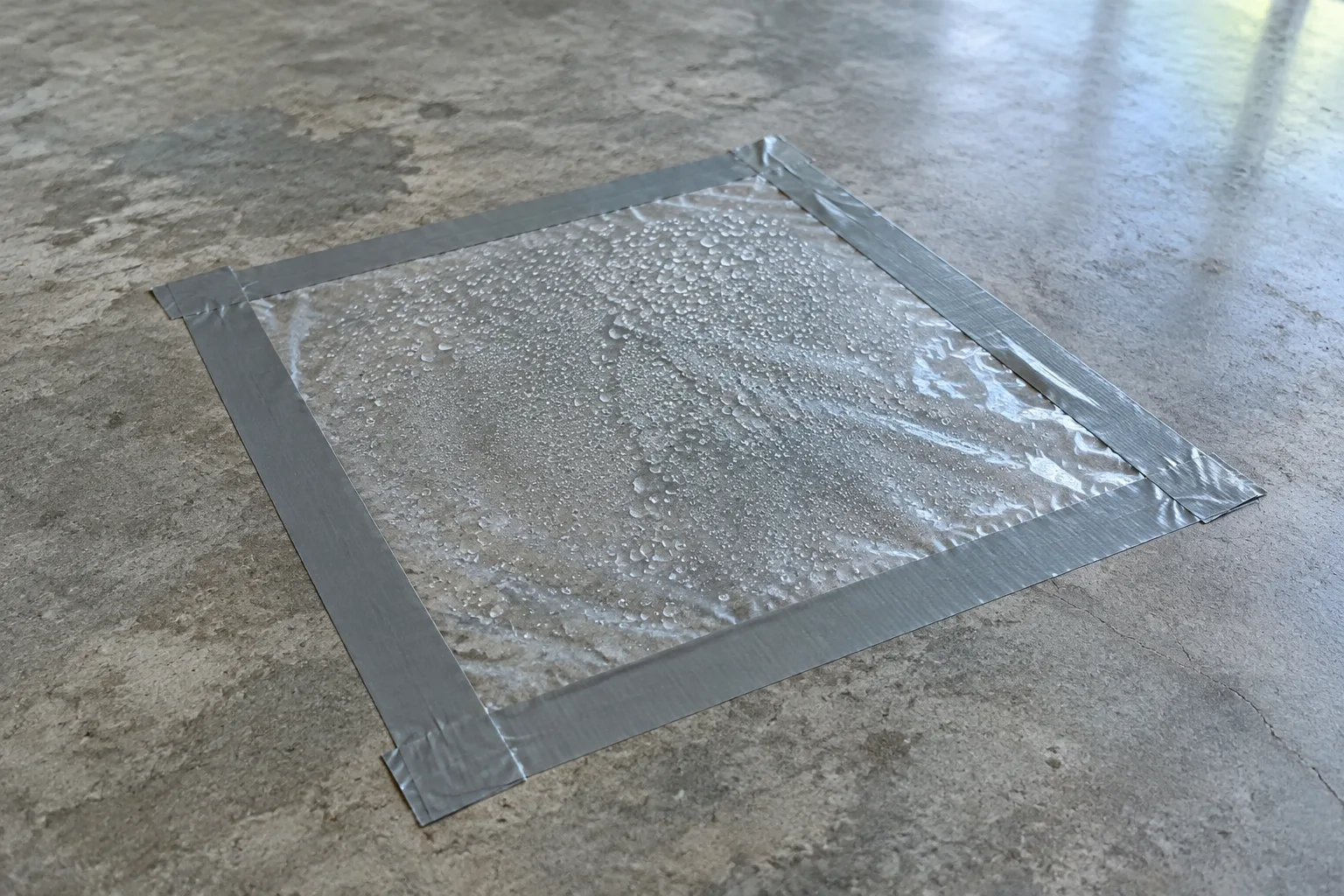 Clear plastic sheet taped down on bare concrete, with condensation droplets collected underneath